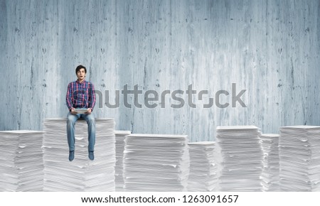 Young man in casual clothing sitting on pile of documents with light-blue wall on background. Mixed media.