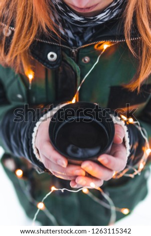 Girl standing near camp fire with a cup of hot coffee in the snowy forest. Concept adventure active vacations outdoor. Winter camping. vertical photo, colorful christmas lights