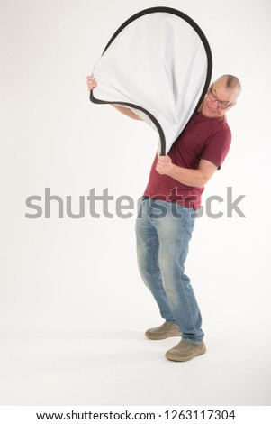 Mature man struggling to put away and fold up a photographic reflector. casual dress in the studio. UK. mature older man.