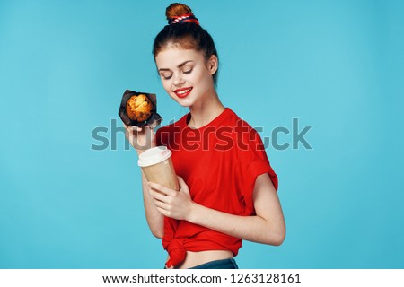 A woman in a red t-shirt holds a cupcake and a cup of drink in her hand                     