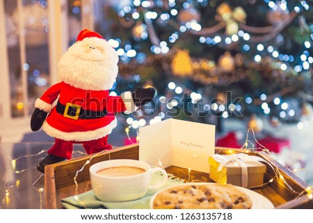 Decorative Santa standing near tray with cocoa, cookies, giftbox and postcard with xmas tree background. Christmas and New Year composition. Festive breakfast. Soft selective focus. Vintage toning