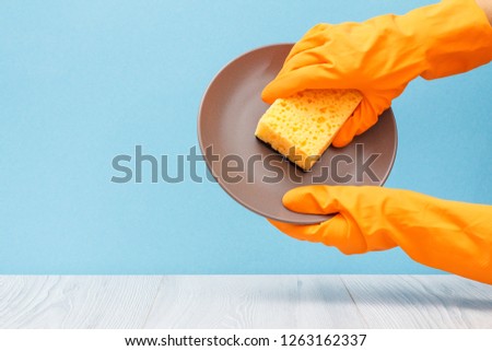 Women's hands in orange protective glove with plate and sponge on blue background. Washing and cleaning concept.