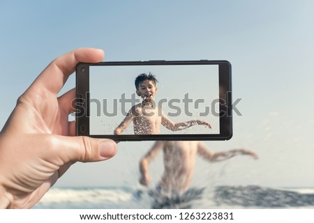 Female taking a picture of a boy on the beach on the phone. Teen boy jumping in sea waves with water splashes. Travel and family concept  