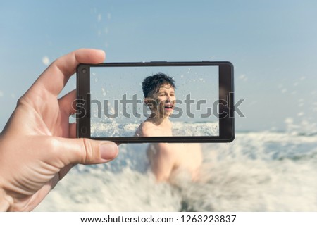 Female taking a picture of a boy on the beach on the phone. Teen boy jumping in sea waves with water splashes. Travel and family concept