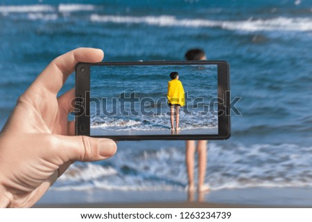 Female taking a picture of a boy in the yellow towel standing on the beach on the phone. Travel and family concept