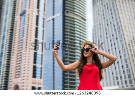 Portrait of young woman in red dress, sunglasses and summer hat make selfie or video call on the phone on downtown skycrapers background