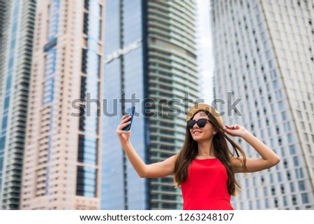 Portrait of young woman in red dress, sunglasses and summer hat take selfie on the phone on downtown skycrapers background