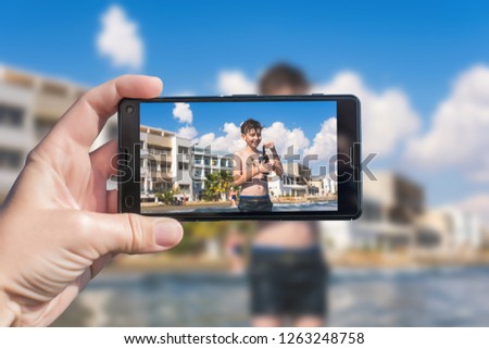 Female taking a picture of a diver boy on the beach on the phone. Happy teen boy in the swimming flippers snorkeling mask and tube.Travel and family concept