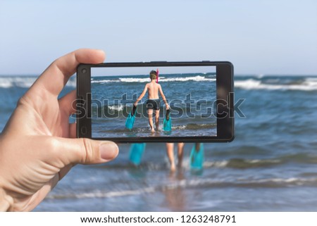 Female taking a picture of a diver boy on the beach on the phone. Happy teen boy in the swimming flippers snorkeling mask and tube.Travel and family concept