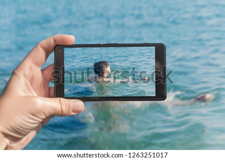 Female taking a picture of diving boy in the sea wave on the phone. Travel and family concept 