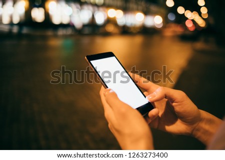 Close-up woman's hands holding mobile phone with blank screen in city at night, streetlights on background.
