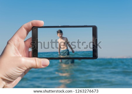 Female taking a picture of a screaming boy on the beach on the phone. Teen boy jumping in sea waves with water splashes. Travel and family concept