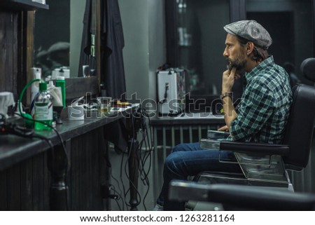 Serious bearded man frowning and touching his chin while looking attentively at his reflection in the mirror
