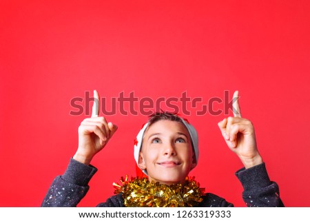 Portrait of a teenager in a Santa hat and with tinsel on his neck, the guy points his fingers at the empty space at the top, on a red background. The concept of Christmas