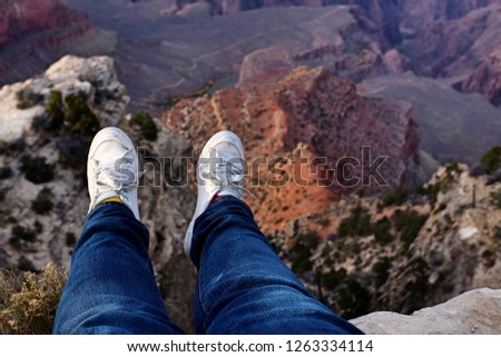 Feet dangle from the edge of a cliff, looking down into a valley in the Grand Canyon