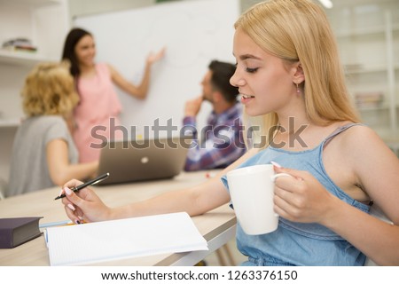Side view of beautiful girl with cup in one hand, sitting at table and writing lecture in notebook. Student wearing in light denim dress, enjoy studying at bright class with group mate on background.