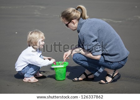 Cute blonde child and her mother playing at the beach with green bucket and pink shovel.