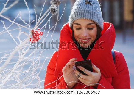 Smilling Girl in red winter jacket types something in her phone standing on the winter street near traditional christmas tree.  Christmas, new year and winter holiday concept - Image