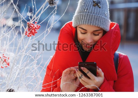 close up portrait of happy woman in red winter down jacket and knitted grey hat types text of sms or message  in her mobile smartphone standing on the winter street near traditional christmas tree. 