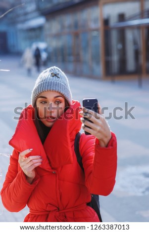 Winter fashion selfie. Young beautiful happy girl in red winter coat and knitted hat and takes selfie in winter street. Christmas, new year and winter holiday concept - Image