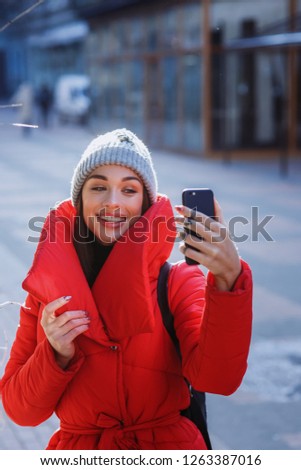 Young happy girl in red down jacket and grey knitted hat takes selfie and play the ape in winter cold day. Girl takes winter selfie. Christmas, new year and winter holiday concept -Image