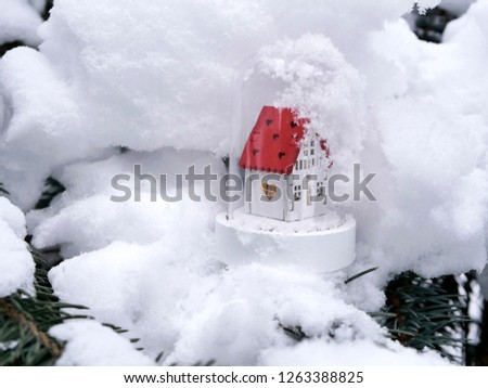 Small toy decorative white house with a red roof and lighted illumination on a snow-covered tree, the concept of winter seasonal holidays, Christmas, a new home in the new year, home comfort
