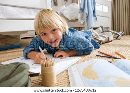 Smiling boy lying on floor in bedroom drawing at camera