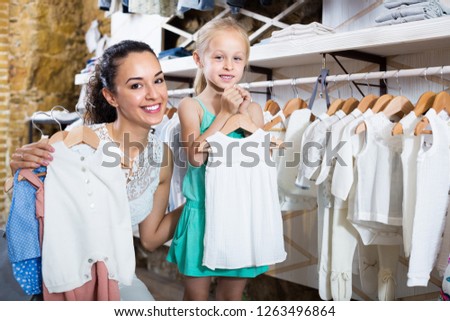Smiling cheerful woman with small girl choosing white baby clothes in kids apparel boutique