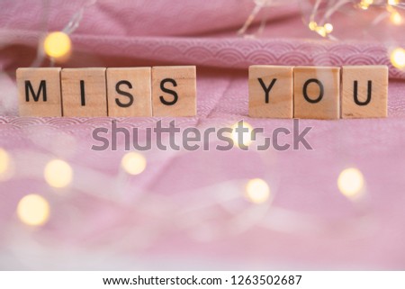 Close up of wooden alphabet letters say Miss You on pink floor decorated with beautiful light bulbs. Image with bokeh in foreground.