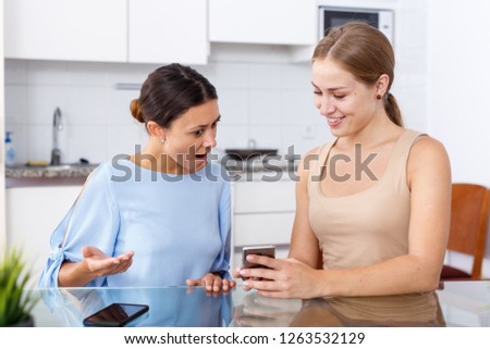 Young smiling girlfriends using phones and chatting at table at home interior