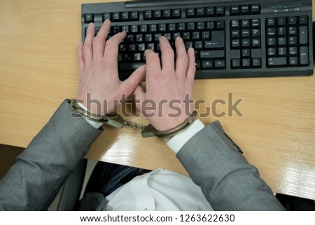 Detail of business person hand tied with handcuffs to workplace, keyboard and monitor in background.