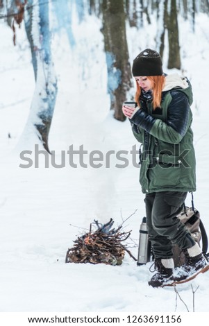 Girl standing near camp fire with a cup of hot coffee in the snowy forest. Concept adventure active vacations outdoor. Winter camping. vertical photo