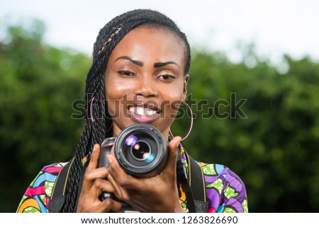 young woman standing right in front of camera with a camera smiling.