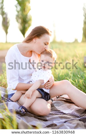 Young blonde woman sitting with little baby on plaid, grass on background. Concept of picnic, motherhood and nature.