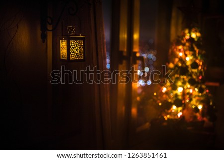 desk of free space and lamp with xmas tree in home . Christmas lantern in selective focus near window with holiday tree full of colored toys and lights. Night scene