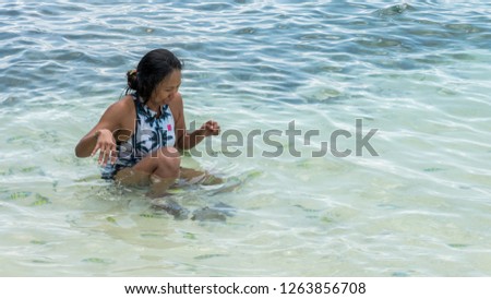 Asian woman sitting in water at beach, laughging and playing while surrounded by tropical fish