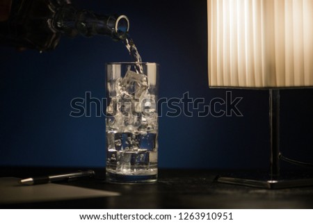 A glass of sparkling water with ice on a dark table with a pen and a sheet of paper and a desk lamp.