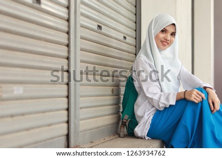 Cute Malaysian girl wearing white school uniform. Girl wearing white hijab. Green backpack. Girl sitting down.