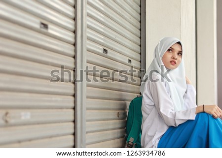 Cute Malaysian girl wearing white school uniform. Girl wearing white hijab. Green backpack. Girl sitting down.
