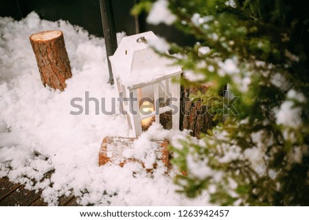 White lamp with candle inside standing on a snowy wooden terrace