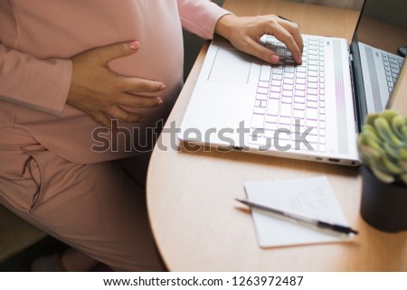 Happy young pregnant woman sitting on bed using technology devices: laptop and cellphone. Future mom expecting child working at home bedroom close-up
