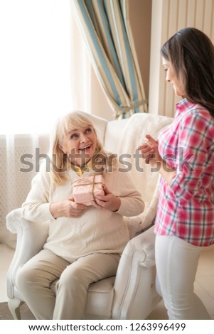 Excited Elderly Woman Sits In The Chair And Looks With Love And Gratitude At Her Charming Daughter While Holding A Present In Pink Wrapping.