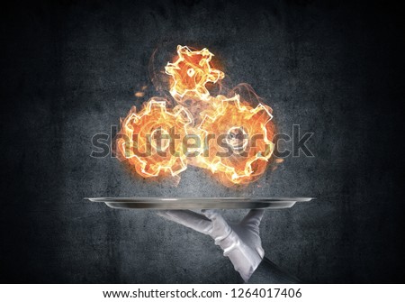 Cropped image of waitress's hand in white glove presenting flaming cogwheels structure on metal tray with dark wall on background.