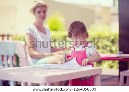 young mother and her little daughter cheerfully spending time together using pencil crayons while drawing a colorful pictures in the outside playschool