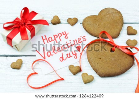 The inscription on the paper" happy Valentine's day ", cookies in the form of hearts and a box with a gift (present) on a white wooden background.