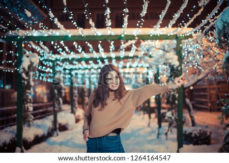 Pretty dark haired girl wearing blue jeans and beige top with snowflakes Christmas lights outdoor at night time.