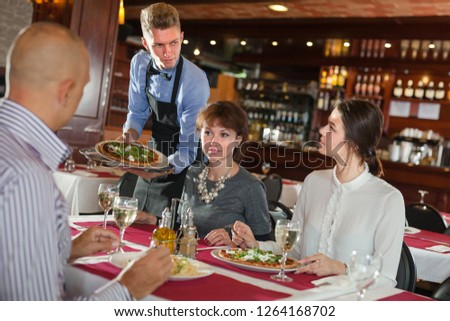 Smiling waiter serving delicious pizza to young visitors of restaurant

