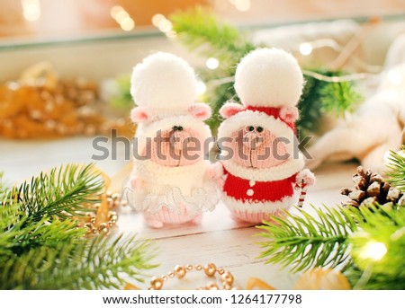 Handmade knitted toys. Christmas pigs in white costume and Santa Claus costume in light christmas background with red berries, branches  and lights