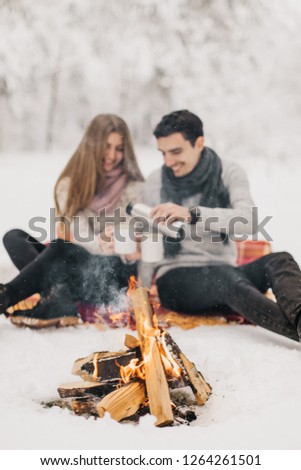 
Young couple on a date in the winter forest is heated by the fire and drink cocoa