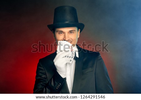 Professional magician wearing suit top hat and gloves standing isolated on blue and red background holding tricks cards posing to camera smiling playful close-up